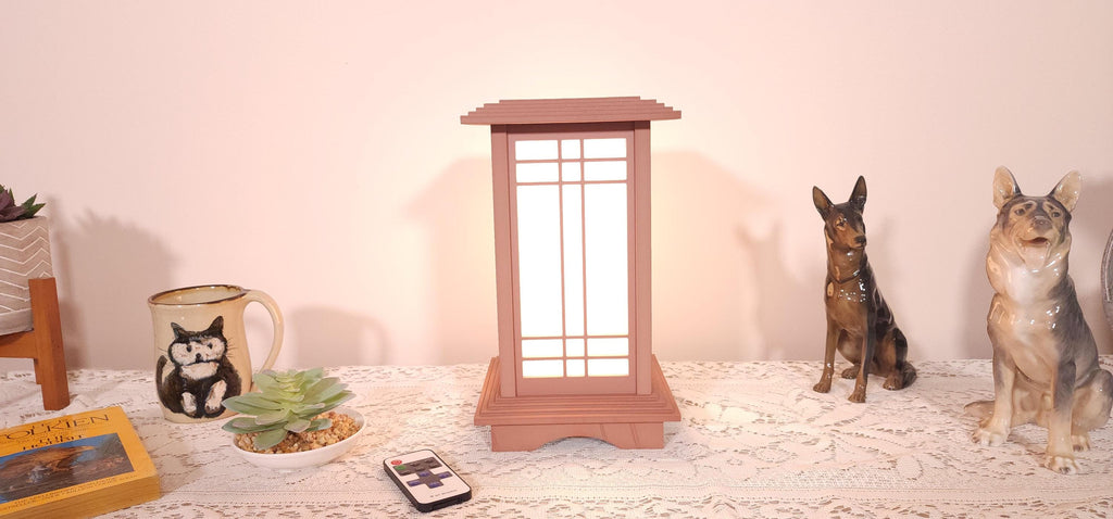 Prairie Beacon Table Lantern in a Cherry wood-look finish, featuring American-inspired architecture, a tiered roof and base, and illuminated geometric lattice panels. The warm white LED lighting shines brightly through four translucent diffusers, creating an inviting display. Shown on a lace-covered table alongside a succulent, ceramic dog figurines, and a small RF remote control for adjusting brightness and lighting modes.