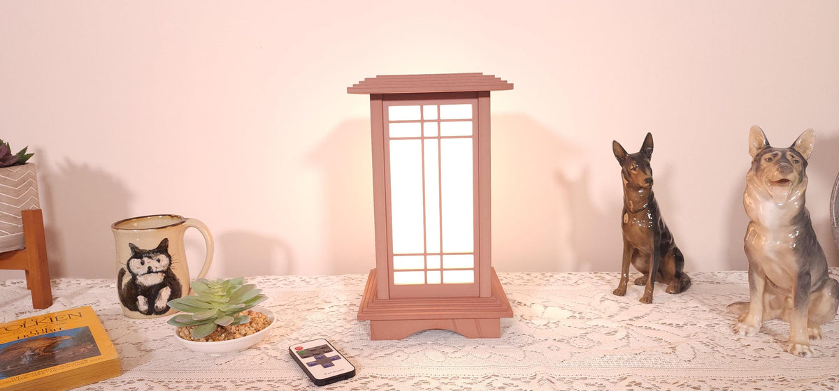 Prairie Beacon Table Lantern in a Cherry wood-look finish, featuring American-inspired architecture, a tiered roof and base, and illuminated geometric lattice panels. The warm white LED lighting shines brightly through four translucent diffusers, creating an inviting display. Shown on a lace-covered table alongside a succulent, ceramic dog figurines, and a small RF remote control for adjusting brightness and lighting modes.