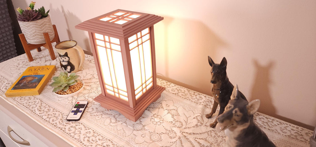 Prairie Beacon Table Lantern in a Cherry wood-look finish, featuring American-inspired architecture, a tiered roof and base, and illuminated geometric lattice panels. The warm white LED lighting shines brightly through four translucent diffusers, creating an inviting display. Shown on a lace-covered table alongside a succulent, ceramic dog figurines, and a small RF remote control for adjusting brightness and lighting modes.