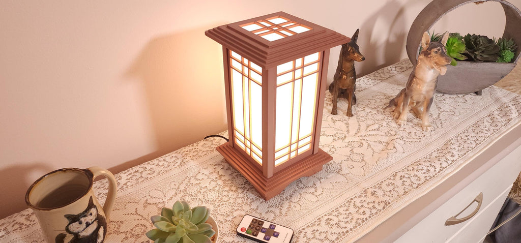 Prairie Beacon Table Lantern in a Cherry wood-look finish, featuring American-inspired architecture, a tiered roof and base, and illuminated geometric lattice panels. The warm white LED lighting shines brightly through four translucent diffusers, creating an inviting display. Shown on a lace-covered table alongside a succulent, ceramic dog figurines, and a small RF remote control for adjusting brightness and lighting modes.