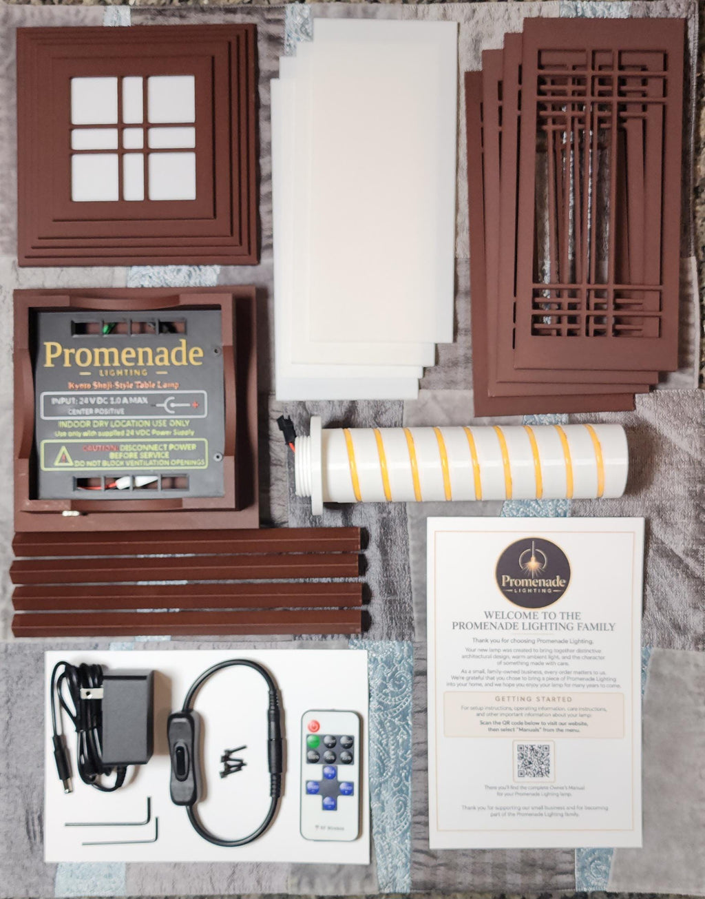 Complete Table Lantern package, with all components arranged before assembly. Shown are the decorative American-inspired wall panels, four translucent white light diffusers, tiered roof and base, four structural posts, and cylindrical LED lighting core. The base features Promenade Lighting branding and electrical safety information. Also included are the 24V power adapter, inline switch, RF remote control, assembly screws, hex keys, and welcome card with QR code linking to the instruction manual.