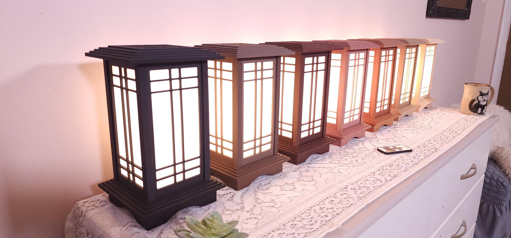 Seven Prairie Beacon Table Lanterns by Promenade Lighting displayed side by side in their complete range of wood-inspired finishes, from deep Ebony and rich Walnut tones to warm Cherry, Teak, Natural Maple, and light Pine. Each lantern features a tiered American-inspired roof and base, geometric architectural window panels, and white light diffusers. The lanterns are arranged on a lace-covered table with a succulent, ceramic cat mug, and RF remote control in front.