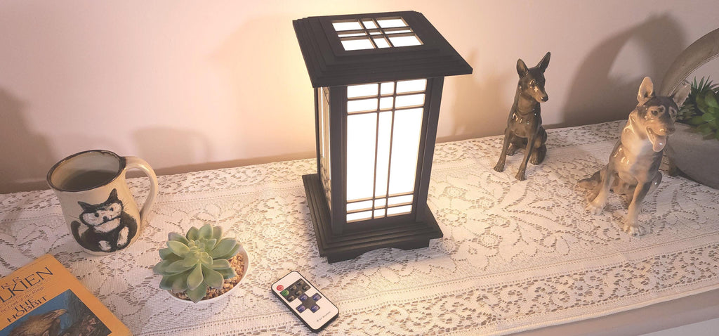 Prairie Beacon Table Lantern in a rich Ebony wood-look finish, featuring American-inspired architecture, a tiered roof and base, and illuminated geometric lattice panels. The warm white LED lighting shines brightly through four translucent diffusers, creating an inviting display. Shown on a lace-covered table alongside a succulent, ceramic dog figurines, and a small RF remote control for adjusting brightness and lighting modes.