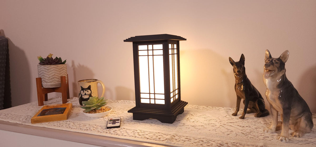 Prairie Beacon Table Lantern in a rich Ebony wood-look finish, featuring American-inspired architecture, a tiered roof and base, and illuminated geometric lattice panels. The warm white LED lighting shines brightly through four translucent diffusers, creating an inviting display. Shown on a lace-covered table alongside a succulent, ceramic dog figurines, and a small RF remote control for adjusting brightness and lighting modes.