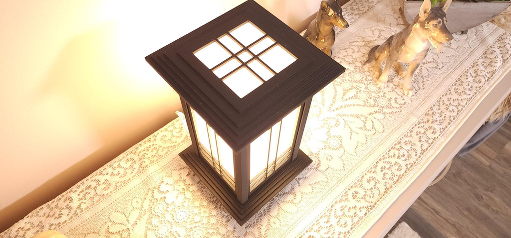 Prairie Beacon Table Lantern in a rich Ebony wood-look finish, featuring American-inspired architecture, a tiered roof and base, and illuminated geometric lattice panels. The warm white LED lighting shines brightly through four translucent diffusers, creating an inviting display. Shown on a lace-covered table alongside a succulent, ceramic dog figurines, and a small RF remote control for adjusting brightness and lighting modes.