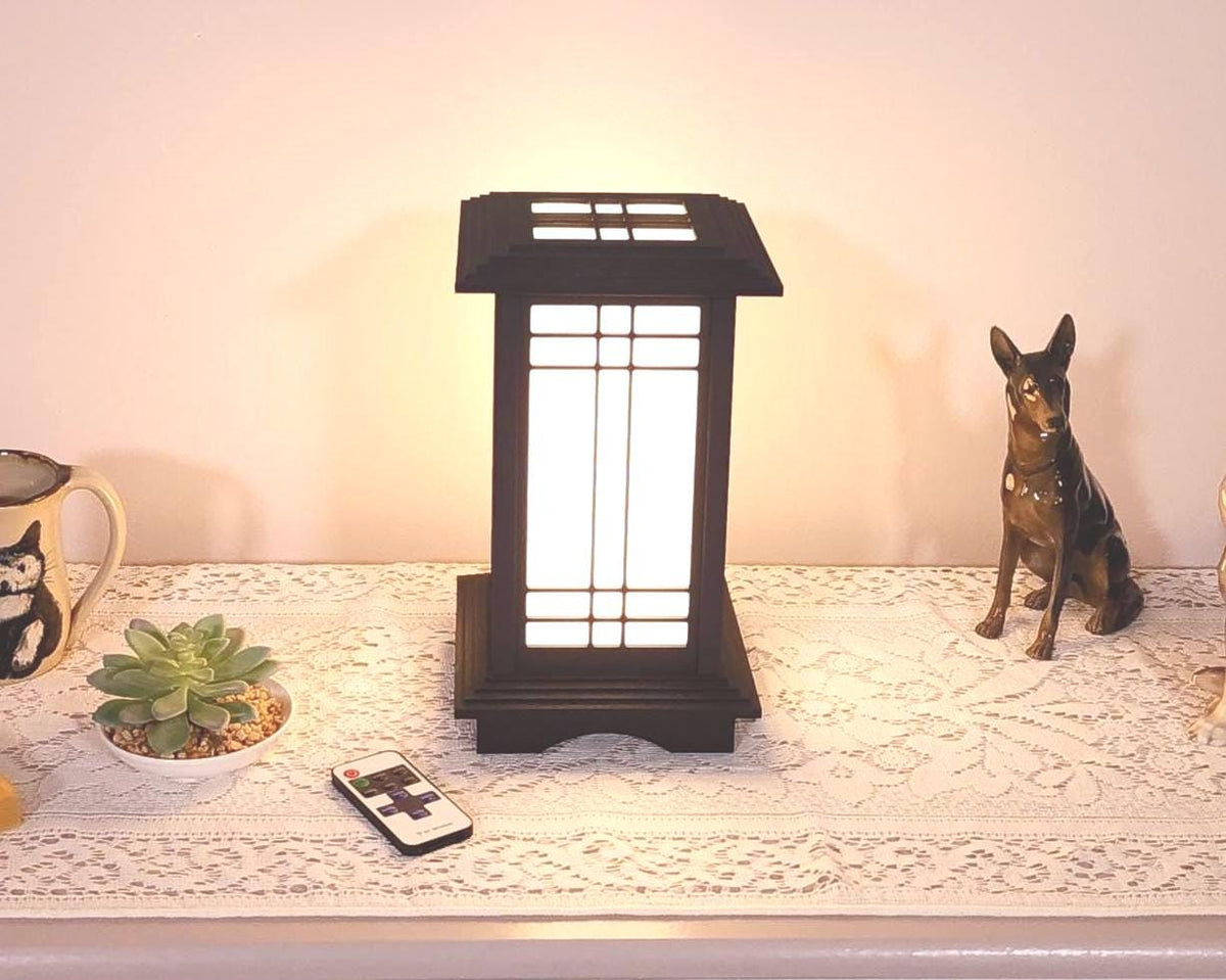 Prairie Beacon Table Lantern in a rich Ebony wood-look finish, featuring American-inspired architecture, a tiered roof and base, and illuminated geometric lattice panels. The warm white LED lighting shines brightly through four translucent diffusers, creating an inviting display. Shown on a lace-covered table alongside a succulent, ceramic dog figurines, and a small RF remote control for adjusting brightness and lighting modes.