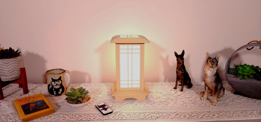 Prairie Beacon Table Lantern in a Maple wood-look finish, featuring American-inspired architecture, a tiered roof and base, and illuminated geometric lattice panels. The warm white LED lighting shines brightly through four translucent diffusers, creating an inviting display. Shown on a lace-covered table alongside a succulent, ceramic dog figurines, and a small RF remote control for adjusting brightness and lighting modes.
