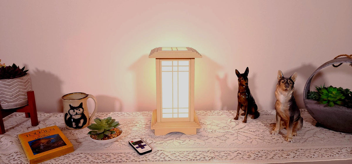 Prairie Beacon Table Lantern in a Maple wood-look finish, featuring American-inspired architecture, a tiered roof and base, and illuminated geometric lattice panels. The warm white LED lighting shines brightly through four translucent diffusers, creating an inviting display. Shown on a lace-covered table alongside a succulent, ceramic dog figurines, and a small RF remote control for adjusting brightness and lighting modes.