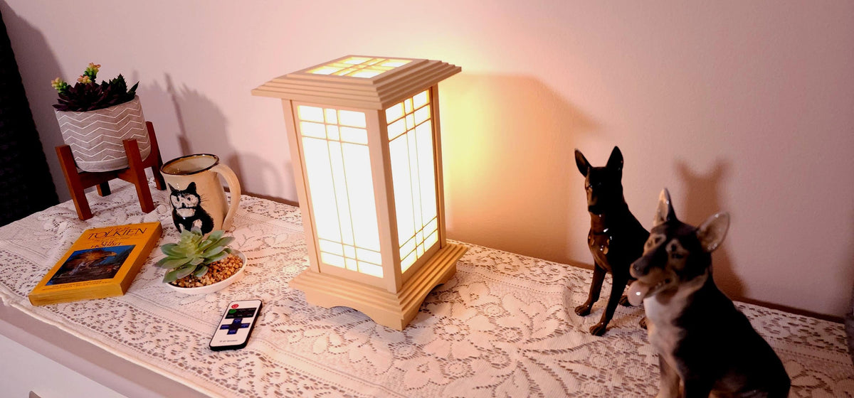 Prairie Beacon Table Lantern in a Maple wood-look finish, featuring American-inspired architecture, a tiered roof and base, and illuminated geometric lattice panels. The warm white LED lighting shines brightly through four translucent diffusers, creating an inviting display. Shown on a lace-covered table alongside a succulent, ceramic dog figurines, and a small RF remote control for adjusting brightness and lighting modes.