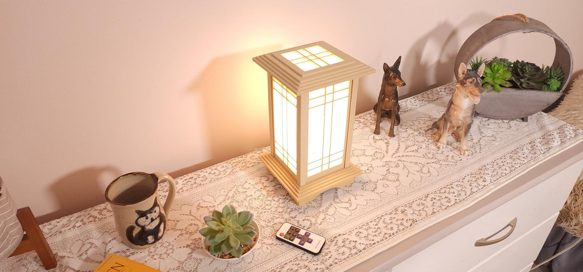 Prairie Beacon Table Lantern in a Maple wood-look finish, featuring American-inspired architecture, a tiered roof and base, and illuminated geometric lattice panels. The warm white LED lighting shines brightly through four translucent diffusers, creating an inviting display. Shown on a lace-covered table alongside a succulent, ceramic dog figurines, and a small RF remote control for adjusting brightness and lighting modes.