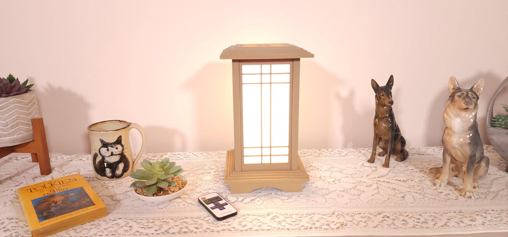 Prairie Beacon Table Lantern in a Pine wood-look finish, featuring American-inspired architecture, a tiered roof and base, and illuminated geometric lattice panels. The warm white LED lighting shines brightly through four translucent diffusers, creating an inviting display. Shown on a lace-covered table alongside a succulent, ceramic dog figurines, and a small RF remote control for adjusting brightness and lighting modes.