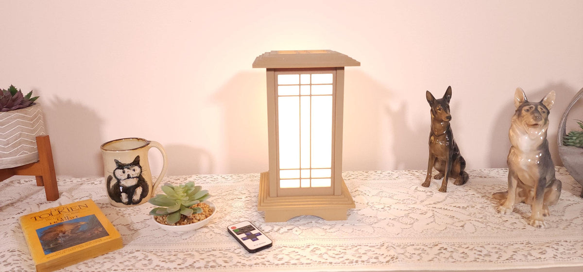 Prairie Beacon Table Lantern in a Pine wood-look finish, featuring American-inspired architecture, a tiered roof and base, and illuminated geometric lattice panels. The warm white LED lighting shines brightly through four translucent diffusers, creating an inviting display. Shown on a lace-covered table alongside a succulent, ceramic dog figurines, and a small RF remote control for adjusting brightness and lighting modes.