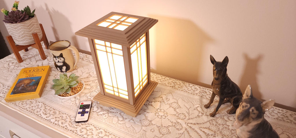 Prairie Beacon Table Lantern in a Pine wood-look finish, featuring American-inspired architecture, a tiered roof and base, and illuminated geometric lattice panels. The warm white LED lighting shines brightly through four translucent diffusers, creating an inviting display. Shown on a lace-covered table alongside a succulent, ceramic dog figurines, and a small RF remote control for adjusting brightness and lighting modes.