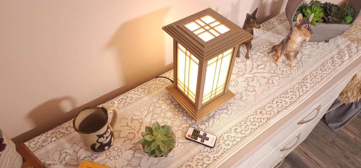 Prairie Beacon Table Lantern in a Pine wood-look finish, featuring American-inspired architecture, a tiered roof and base, and illuminated geometric lattice panels. The warm white LED lighting shines brightly through four translucent diffusers, creating an inviting display. Shown on a lace-covered table alongside a succulent, ceramic dog figurines, and a small RF remote control for adjusting brightness and lighting modes.