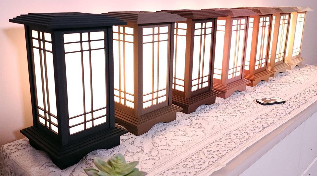 Seven Prairie Beacon Table Lanterns by Promenade Lighting displayed side by side in their complete range of wood-inspired finishes, from deep Ebony and rich Walnut tones to warm Cherry, Teak, Natural Maple, and light Pine. Each lantern features a tiered American-inspired roof and base, geometric architectural window panels, and white light diffusers. The lanterns are arranged on a lace-covered table with a succulent, ceramic cat mug, and RF remote control in front.