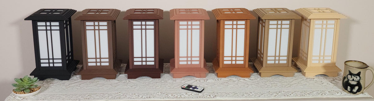 Seven Prairie Beacon Table Lanterns by Promenade Lighting displayed side by side in their complete range of wood-inspired finishes, from deep Ebony and rich Walnut tones to warm Cherry, Teak, Natural Maple, and light Pine. Each lantern features a tiered American-inspired roof and base, geometric architectural window panels, and white light diffusers. The lanterns are arranged on a lace-covered table with a succulent, ceramic cat mug, and RF remote control in front.