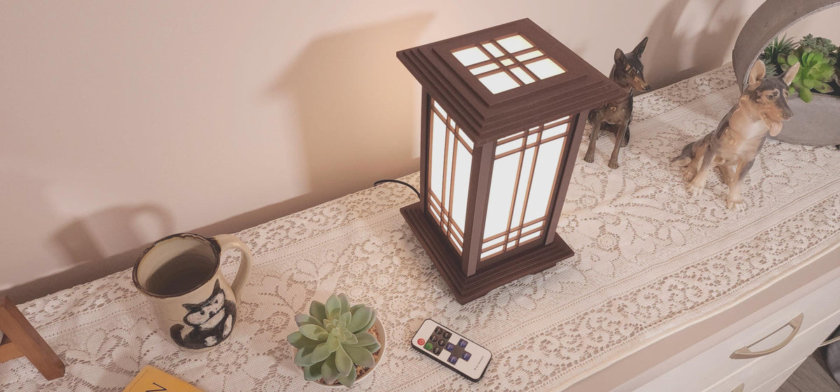 Prairie Beacon Table Lantern in a Rosewood wood-look finish, featuring American-inspired architecture, a tiered roof and base, and illuminated geometric lattice panels. The warm white LED lighting shines brightly through four translucent diffusers, creating an inviting display. Shown on a lace-covered table alongside a succulent, ceramic dog figurines, and a small RF remote control for adjusting brightness and lighting modes.