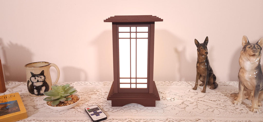Prairie Beacon Table Lantern in a Rosewood wood-look finish, featuring American-inspired architecture, a tiered roof and base, and illuminated geometric lattice panels. The warm white LED lighting shines brightly through four translucent diffusers, creating an inviting display. Shown on a lace-covered table alongside a succulent, ceramic dog figurines, and a small RF remote control for adjusting brightness and lighting modes.