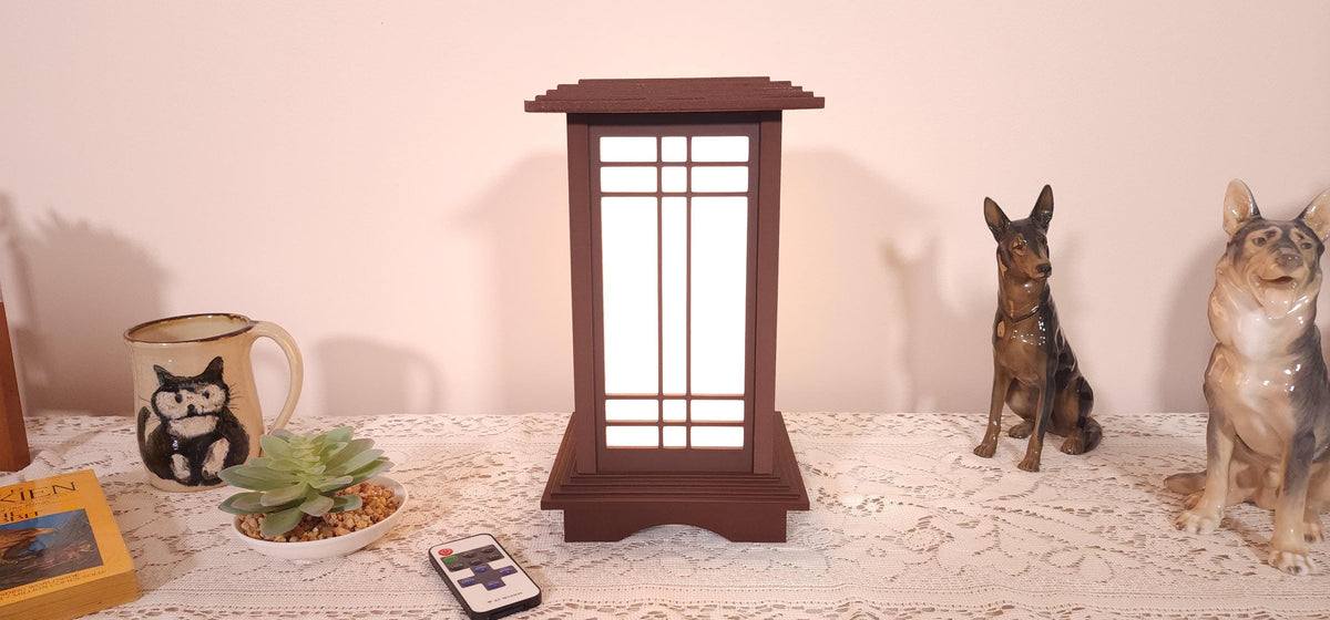 Prairie Beacon Table Lantern in a Rosewood wood-look finish, featuring American-inspired architecture, a tiered roof and base, and illuminated geometric lattice panels. The warm white LED lighting shines brightly through four translucent diffusers, creating an inviting display. Shown on a lace-covered table alongside a succulent, ceramic dog figurines, and a small RF remote control for adjusting brightness and lighting modes.