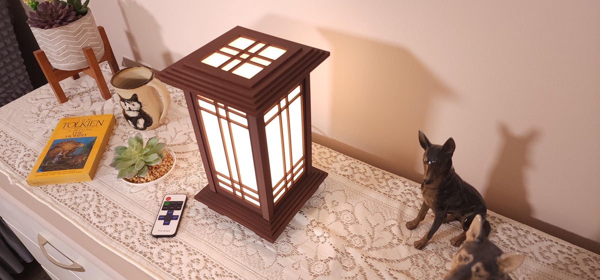 Prairie Beacon Table Lantern in a Rosewood wood-look finish, featuring American-inspired architecture, a tiered roof and base, and illuminated geometric lattice panels. The warm white LED lighting shines brightly through four translucent diffusers, creating an inviting display. Shown on a lace-covered table alongside a succulent, ceramic dog figurines, and a small RF remote control for adjusting brightness and lighting modes.