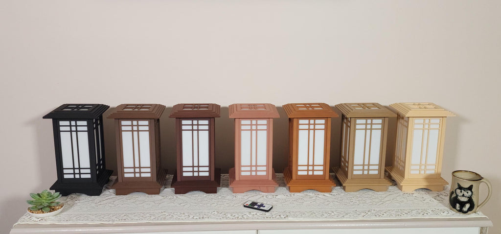 Seven Prairie Beacon Table Lanterns by Promenade Lighting displayed side by side in their complete range of wood-inspired finishes, from deep Ebony and rich Walnut tones to warm Cherry, Teak, Natural Maple, and light Pine. Each 11-inch lantern features a tiered American-inspired roof and base, geometric architectural window panels, and white light diffusers. The lanterns are arranged on a lace-covered table with a succulent, ceramic cat mug, and RF remote control in front.