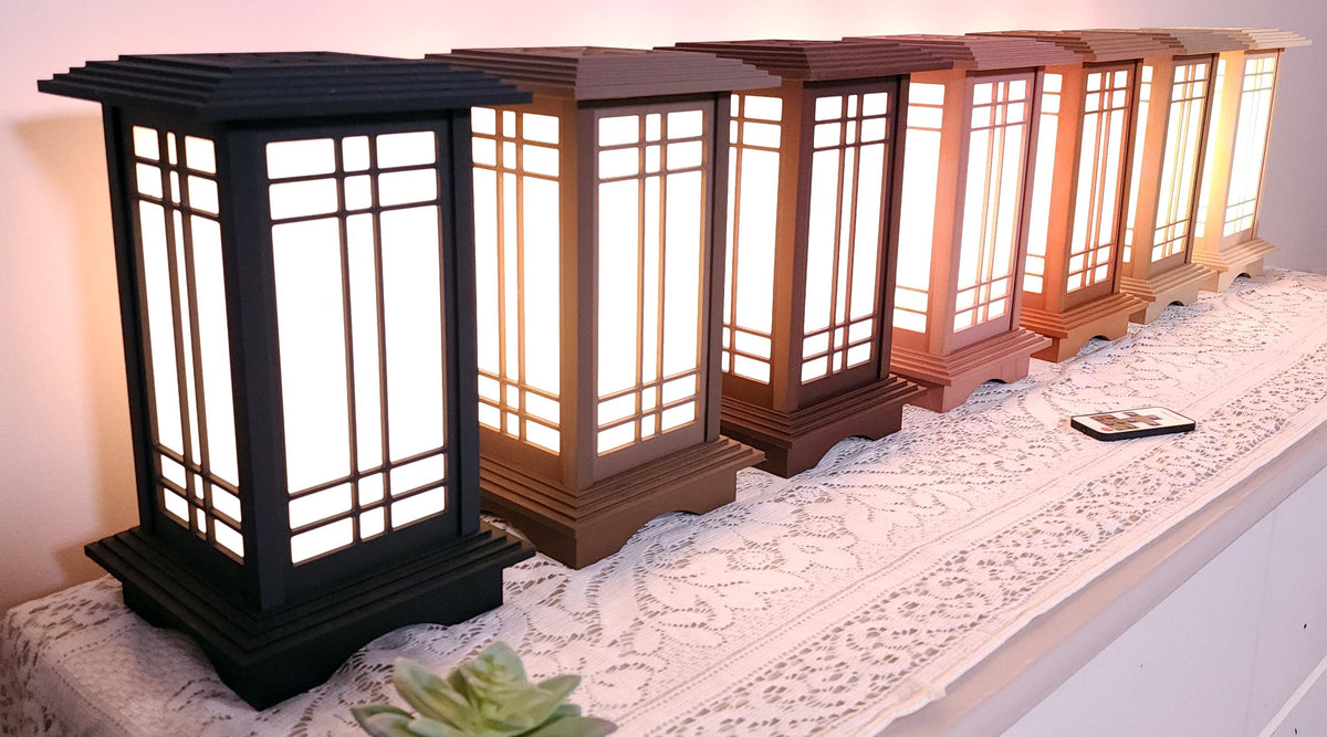 Seven Prairie Beacon Table Lanterns by Promenade Lighting displayed side by side in their complete range of wood-inspired finishes, from deep Ebony and rich Walnut tones to warm Cherry, Teak, Natural Maple, and light Pine. Each 11-inch lantern features a tiered American-inspired roof and base, geometric architectural window panels, and white light diffusers. The lanterns are arranged on a lace-covered table with a succulent, ceramic cat mug, and RF remote control in front.