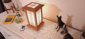 Prairie Beacon Table Lantern in a Teak wood-look finish, featuring American-inspired architecture, a tiered roof and base, and illuminated geometric lattice panels. The warm white LED lighting shines brightly through four translucent diffusers, creating an inviting display. Shown on a lace-covered table alongside a succulent, ceramic dog figurines, and a small RF remote control for adjusting brightness and lighting modes.
