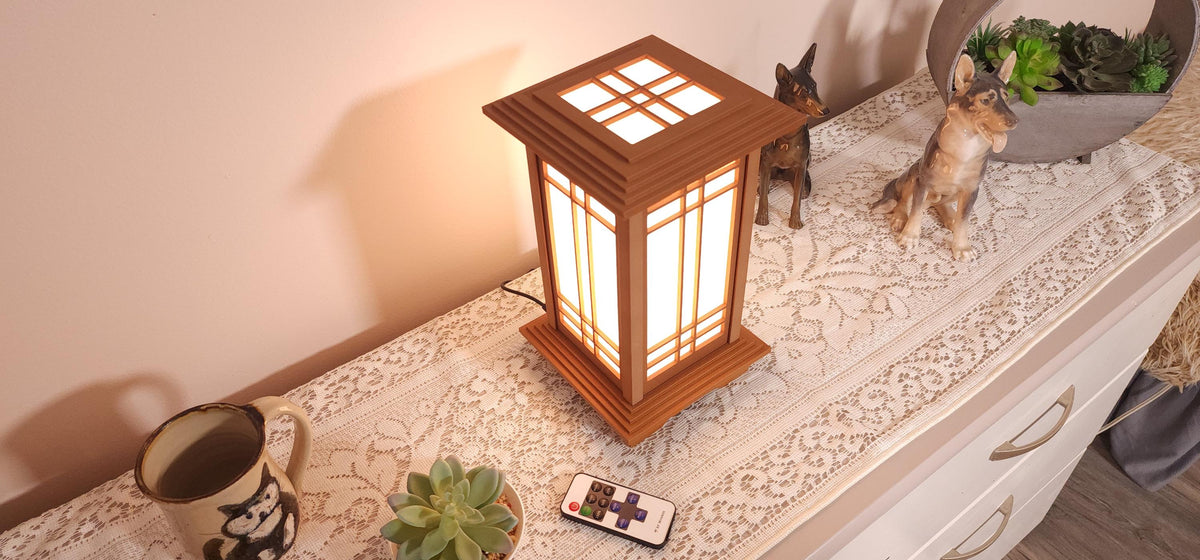 The Prairie Beacon table lantern pairs an inviting Teak wood-tone finish with the sculptural elegance of American-inspired architecture. Slender corner posts, intricate geometric latticework, and translucent panels rise from a square base to a gracefully overhanging roof. Thoughtfully designed and 3D printed, its layered details create a distinctive statement piece, bringing warmth, character, and architectural beauty to a bedside table, bookshelf, or favorite corner.