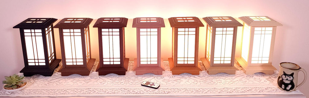 Seven Prairie Beacon Table Lanterns by Promenade Lighting displayed side by side in their complete range of wood-inspired finishes, from deep Ebony and rich Walnut tones to warm Cherry, Teak, Natural Maple, and light Pine. Each lantern features a tiered American-inspired roof and base, geometric architectural window panels, and white light diffusers. The lanterns are arranged on a lace-covered table with a succulent, ceramic cat mug, and RF remote control in front.