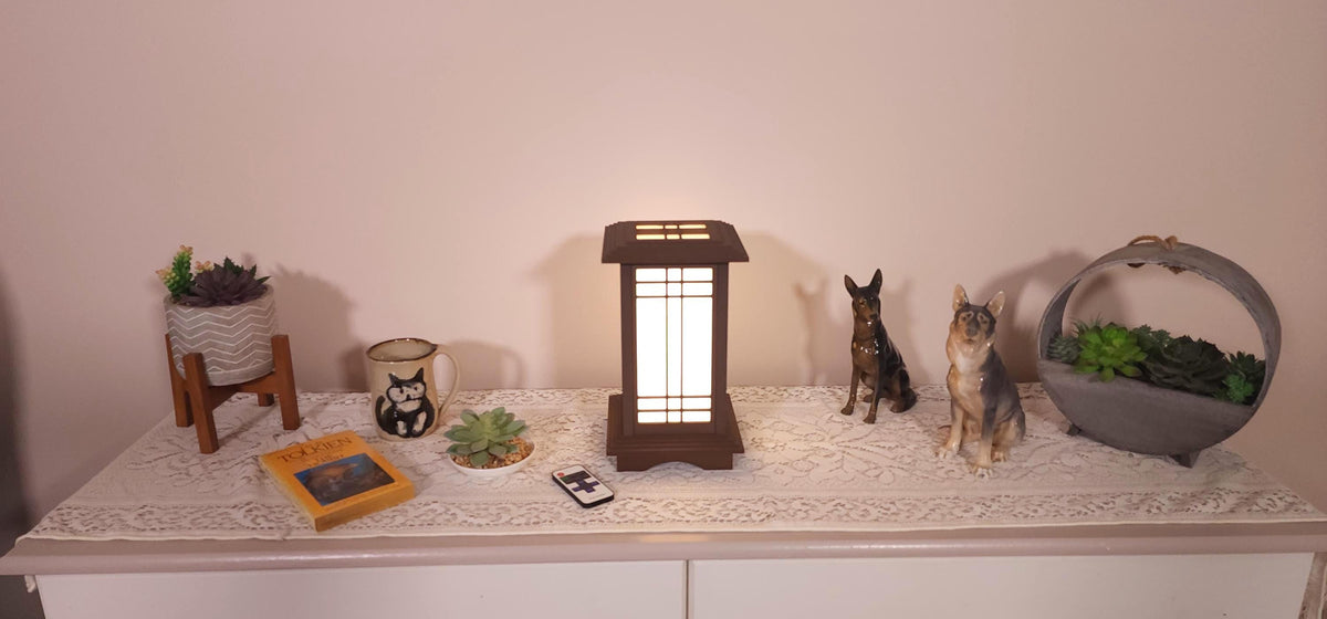 Prairie Beacon Table Lantern in a rich Mission Walnut wood-look finish, featuring American-inspired architecture, a tiered roof and base, and illuminated geometric lattice panels. The warm white LED lighting shines brightly through four translucent diffusers, creating an inviting display. Shown on a lace-covered table alongside a succulent, ceramic dog figurines, and a small RF remote control for adjusting brightness and lighting modes. Complete lantern by Promenade Lighting.