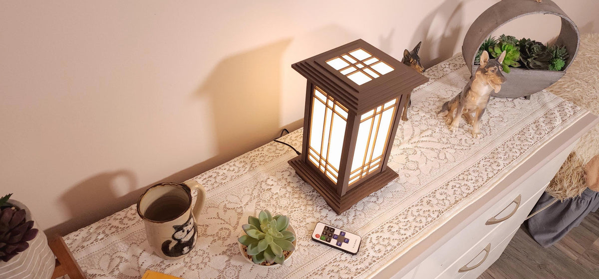 Prairie Beacon Table Lantern in a rich Mission Walnut wood-look finish, featuring American-inspired architecture, a tiered roof and base, and illuminated geometric lattice panels. The warm white LED lighting shines brightly through four translucent diffusers, creating an inviting display. Shown on a lace-covered table alongside a succulent, ceramic dog figurines, and a small RF remote control for adjusting brightness and lighting modes.