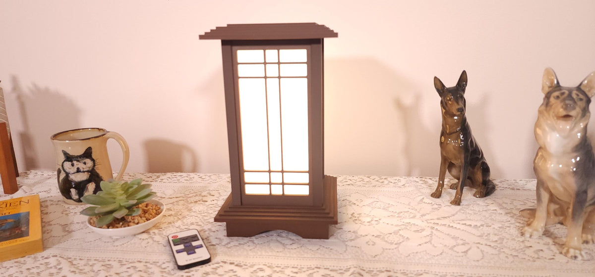 Prairie Beacon Table Lantern in a rich Mission Walnut wood-look finish, featuring American-inspired architecture, a tiered roof and base, and illuminated geometric lattice panels. The warm white LED lighting shines brightly through four translucent diffusers, creating an inviting display. Shown on a lace-covered table alongside a succulent, ceramic dog figurines, and a small RF remote control for adjusting brightness and lighting modes.